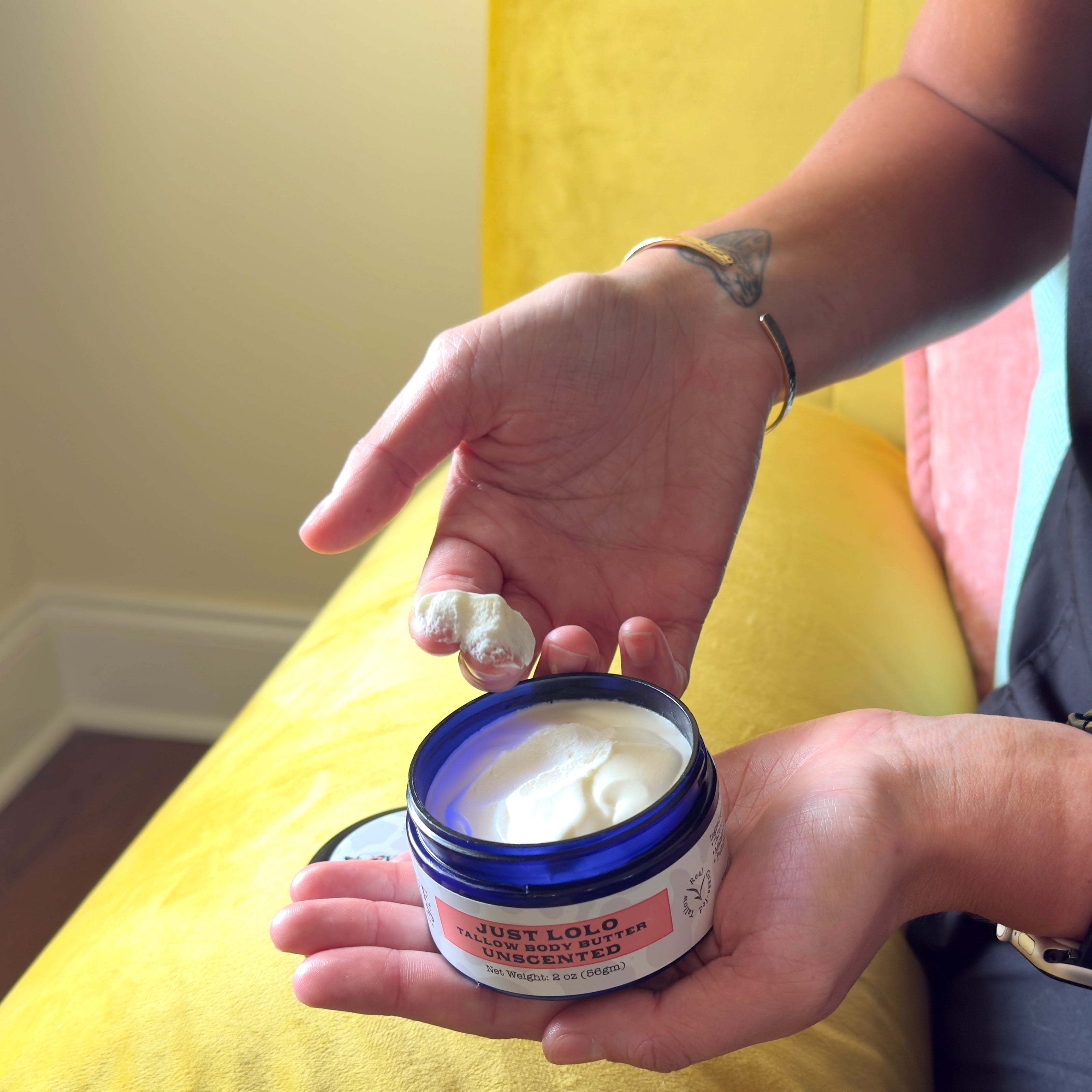 Person holding a jar of cream with a small amount on their finger, sitting on a yellow couch.
