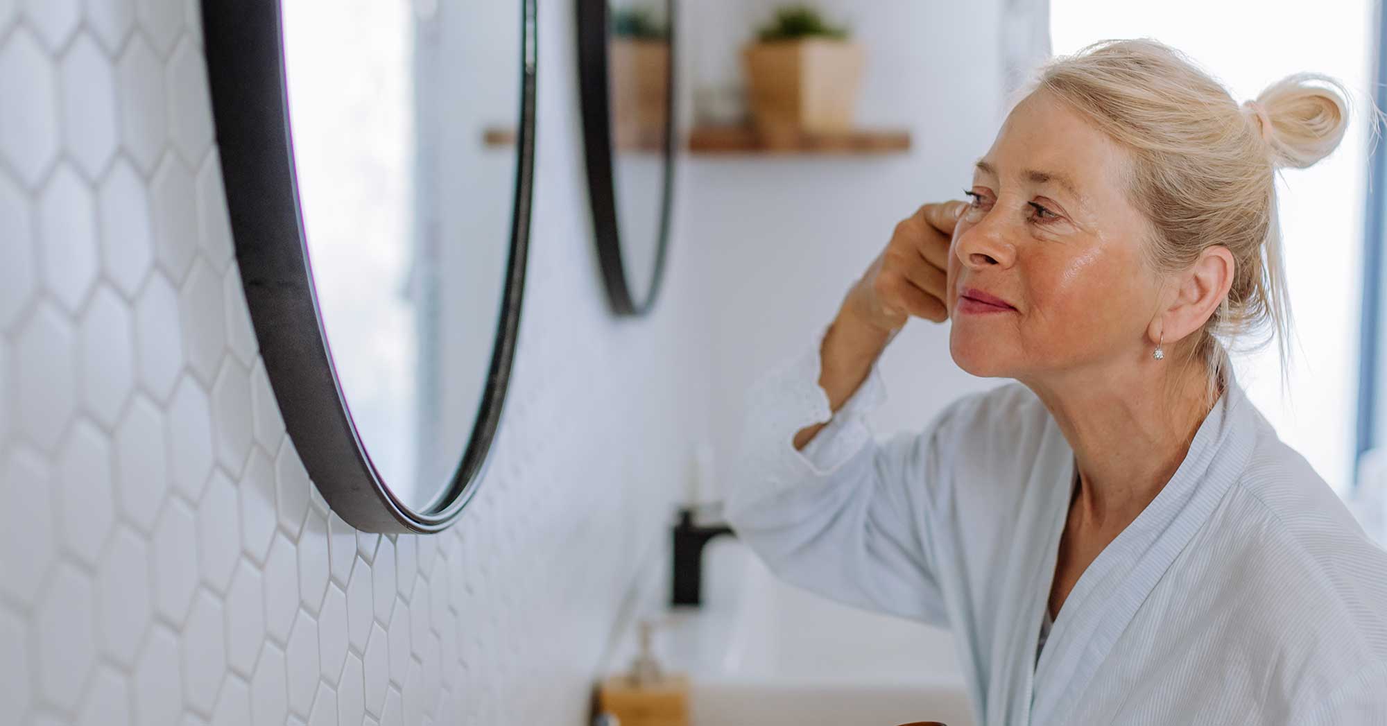 Woman in a white robe looking at herself in a bathroom mirror.