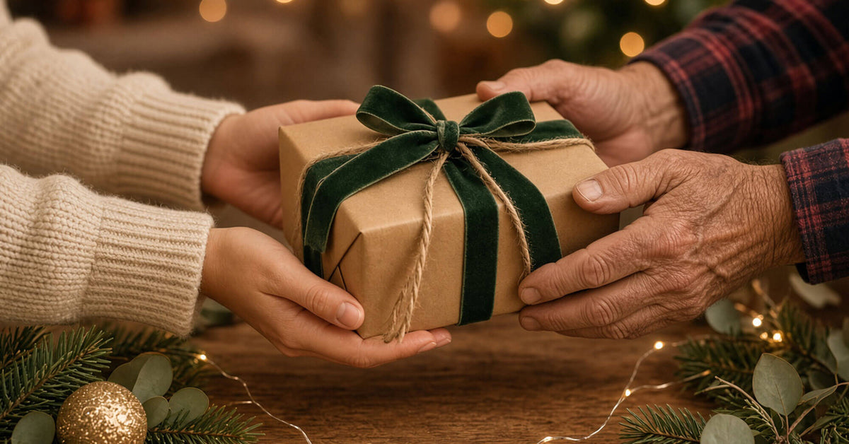 A person in a cream knit sweater hands a brown kraft paper gift box tied with a dark green velvet ribbon and bow to another person wearing a red plaid shirt. The scene is decorated with pine branches, fairy lights, and a gold ornament on a wooden table.