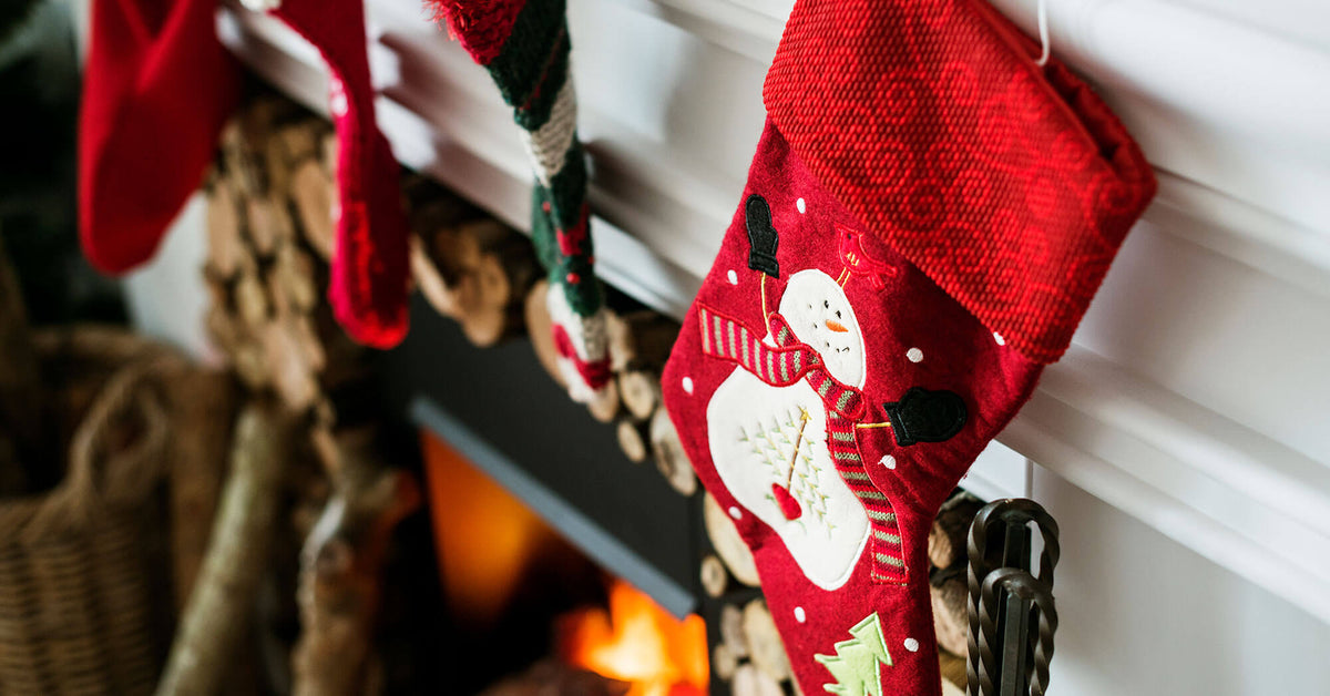 A red Christmas stocking with a white snowman design hangs from a white fireplace mantel above a burning fire with logs visible in the background.