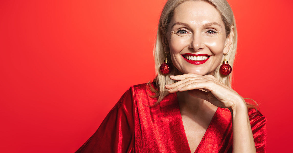 A smiling blonde woman with red lipstick and red spherical earrings, wearing a shiny red satin top, posed against a solid red background.